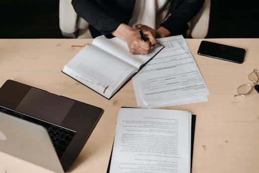 Overhead view of a desk setup with business documents, a laptop, and a person writing.